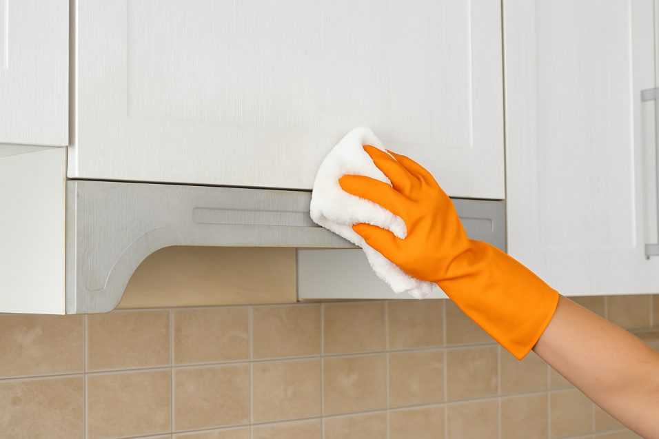 Mano con guanto arancione che pulisce un pensile della cucina con un panno in microfibra, rimuovendo lo sporco e il grasso accumulato