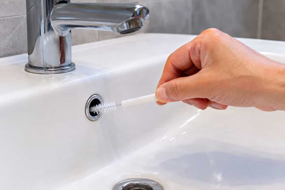 Mano che pulisce con una piccola spazzola il foro del troppo pieno nella parte alta di un lavabo del bagno.
